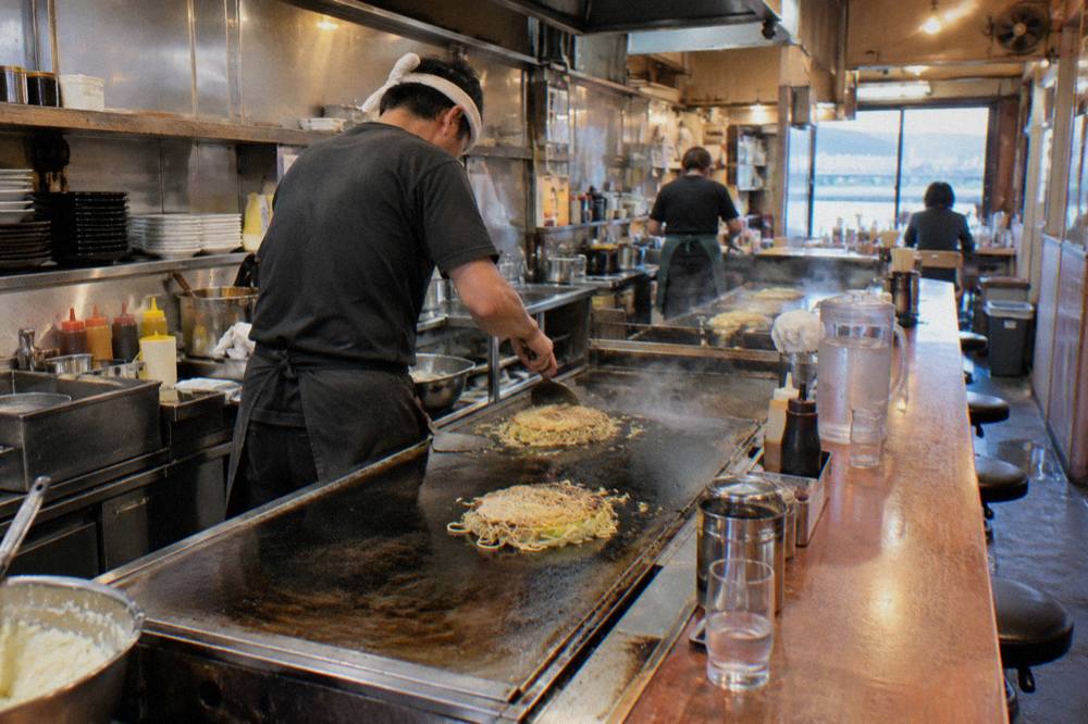 A narrow okonomiyaki counter restaurant with a chef working two teppan griddles