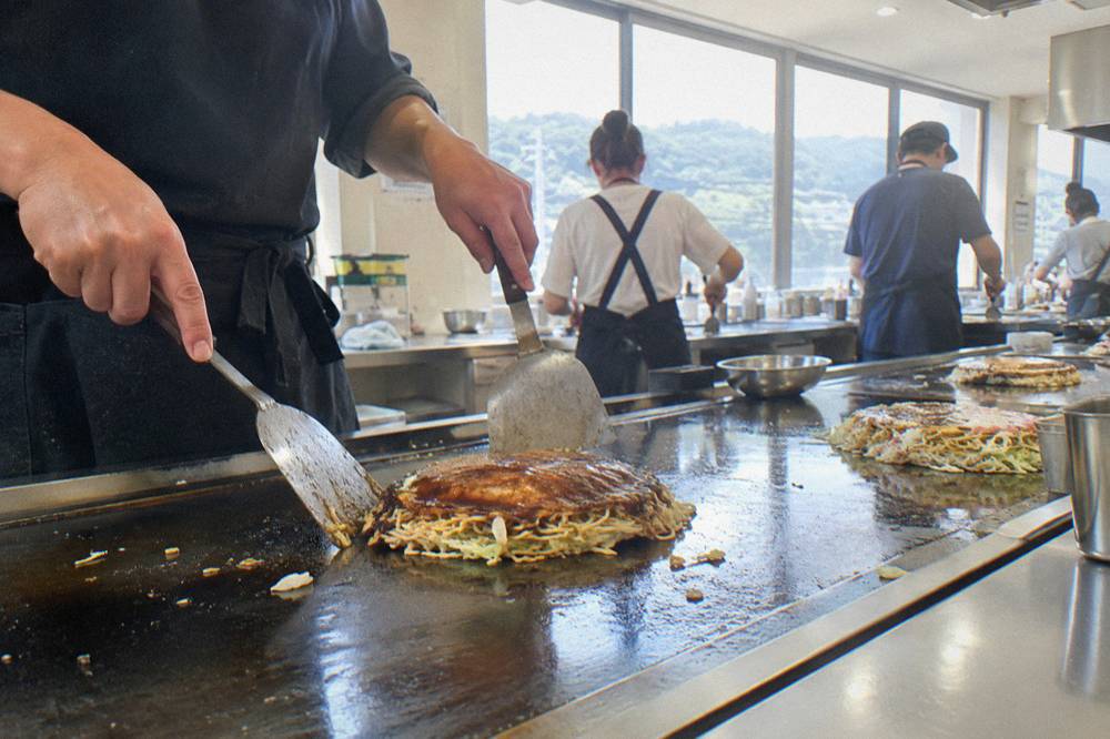 Two spatulas turning a layered okonomiyaki stack on a hot griddle